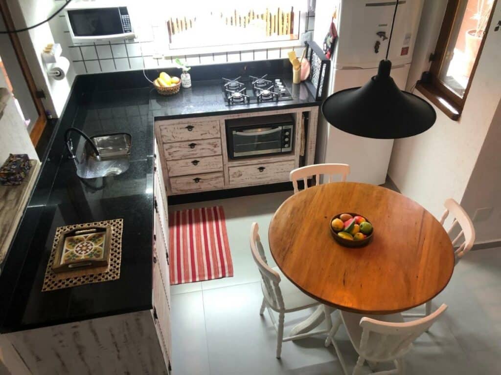 Cozinha do Chalé Boiçucanga. O balcão com pia, fogão e mini forno ocupam duas paredes. Ao lado está uma geladeira. No meio do ambiente há uma mesa redonda de quatro lugares. Essa imagem faz parte dos airbnb em Boiçucanga.