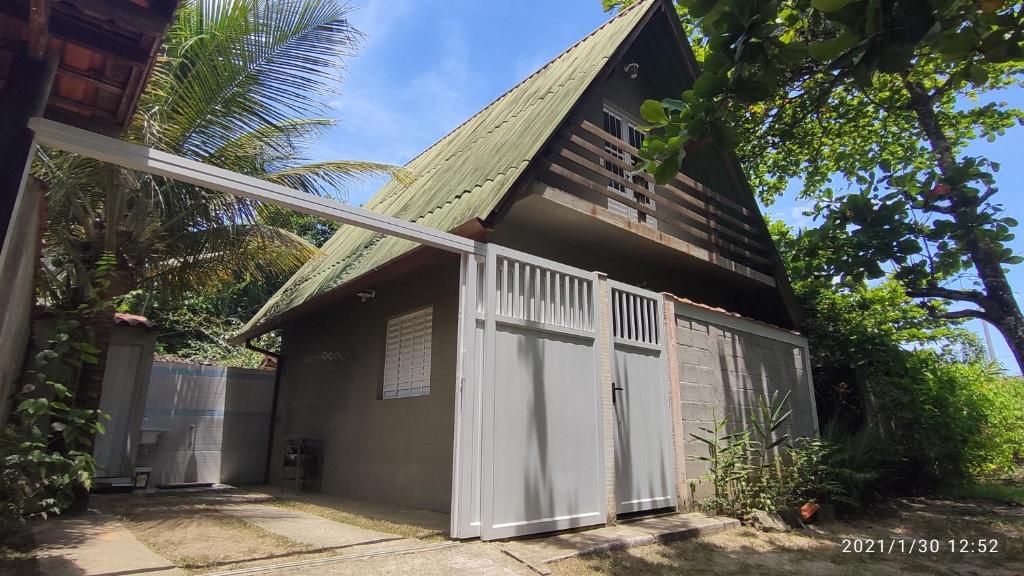 Fachada do Chale da Dalva que mostra um portão de correr aberto que dá acesso a garagem do chalé com uma palmeira e do lado a construção do chalé com telhado pontudo. Imagem ilustrando post Airbnb em Picinguaba.
