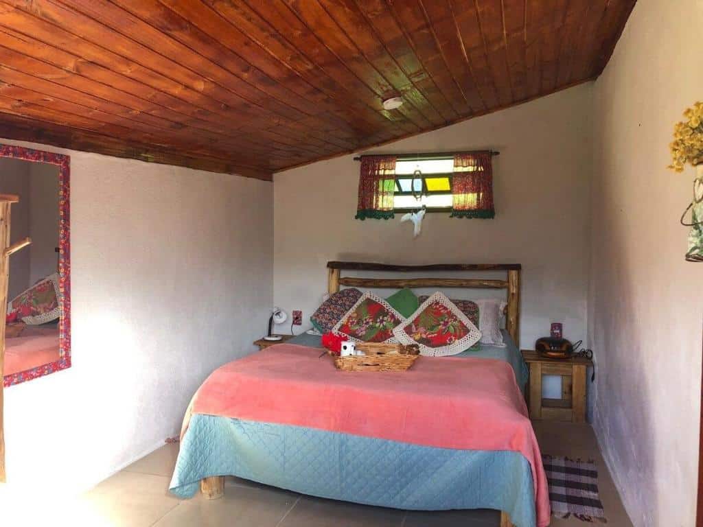Imagem do quarto em Chalé jeito de roça na Pedra Selada, que está ilustrando o post sobre airbnb em Visconde de Mauá. Há uma cama de casal entre duas mesas de cabeceira. Na esquerda há um espelho. 