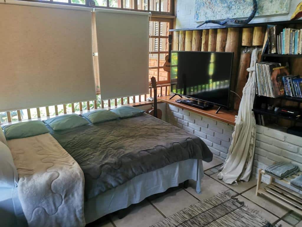 Imagem do quarto em Chalé Mauá Conforto e Arte, que está ilustrando o post sobre airbnb em Visconde de Mauá. A cama de casal está na direita e vemos ela de lado, e do outro lado dela há uma janela com persianas. Na frente há uma TV e estantes com livros, além de um violão encostado no pé da cama.