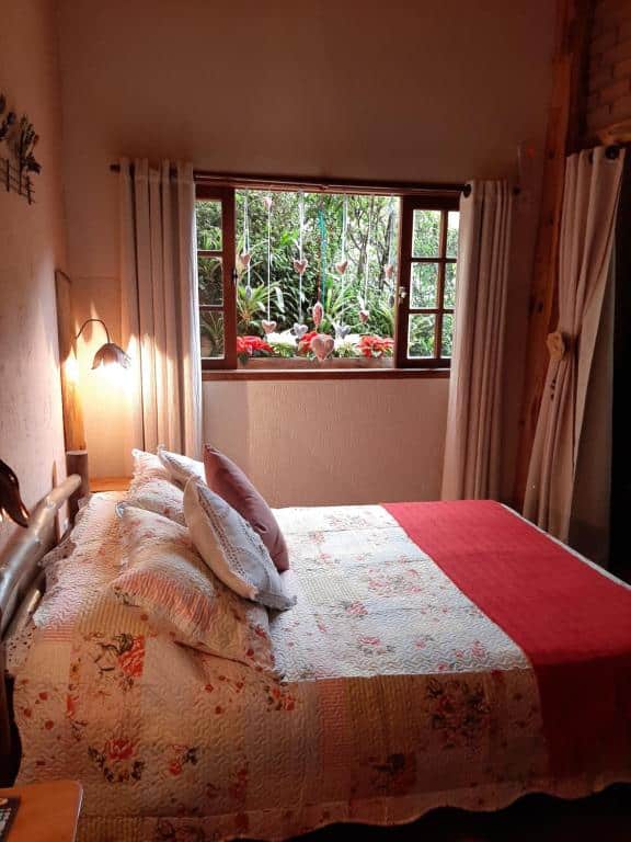 Imagem do quarto em Chalé Raio De Luz, que está ilustrando o post sobre airbnb em Visconde de Mauá. Vemos a cama de lado, e é de casal, e do outro lado dela há uma janela com decorações românticas e vista para um jardim.