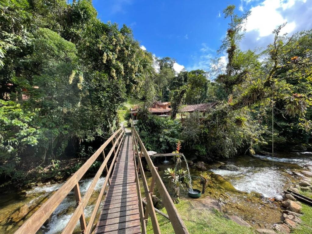 Imagem na natureza no Chalé Truta da Floresta. Há uma ponte que passa por cima de uma cachoeira, e em volt há várias árvores e os chalés estão no fundo.