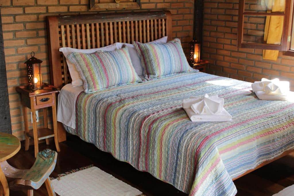 Imagem do quarto em Chalés de Analuz, que está ilustrando o post sobre airbnb em Visconde de Mauá. As paredes são de tijolos e há uma cama de casal no meio da foto. Na direita da cama há uma janela, e de seus dois lados há uma mesa de cabeceira com abajur.
