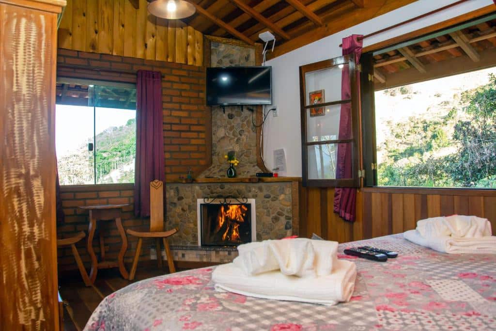 Imagem do quarto em Chalés Encanto do sol, que está ilustrando o post sobre airbnb em Visconde de Mauá. Vemos a cama de relance, e na frente dela há uma lareira e TV entre duas janelas.