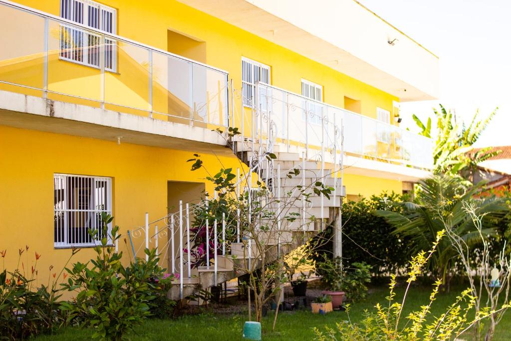 Parte dos Chalés Mallumar que mostra no térreo um jardim verde com plantas e flores e uma escada em formato caracol que sobe para o segundo andar da construção amarela do chalé. Imagem ilustrando post Airbnb em Maranduba.
