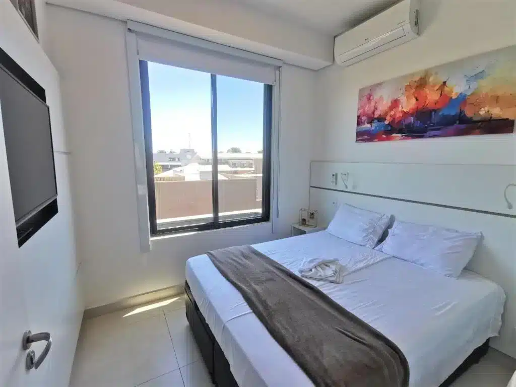 Quarto do Click Apê Cataratas Luxo. Há uma cama box de casala na direita, uma cabeceira atrás e um quadro. Um ar-condicionado está no topo da parede. Uma janela está à direta da cama. Uma TV está na frente da cama.