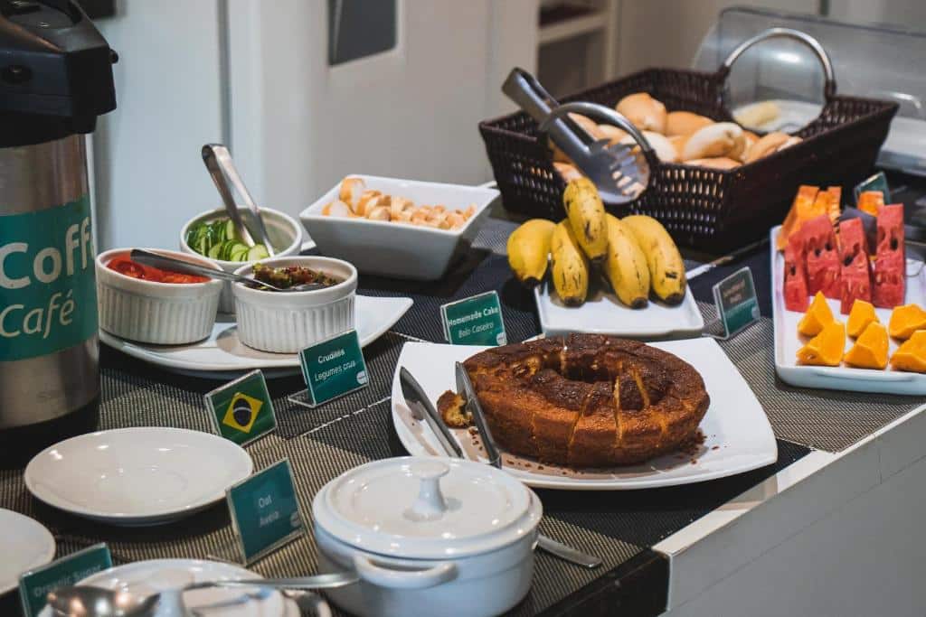Foto do Concept Design Hostel & Suítes, mostrando um balcão com pratos de porcelana com diversos alimentos etiquetados, como bolo, frutas, pães, café, etc.