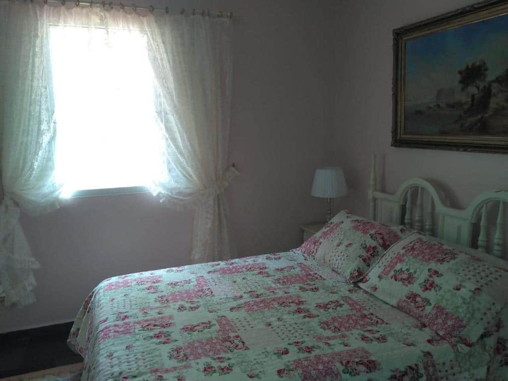 Foto do quarto em Condomínio Vila Paradiso. As paredes são rosa claro. Há uma cama no centro com cabeceira de madeira branca, e ao seu lado na direita há um gaveteiro branco de madeira. Há um quadro atrás da cama, decorando a parede. 
Ilustra o post sobre airbnb em Serra Negra.