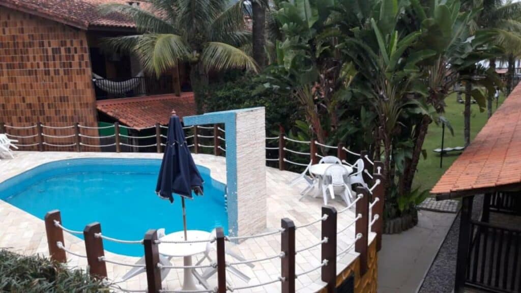 Piscina do Condominio Frente Praia Boissucanga. Ela está em uma área alta com algumas mesas, cadeiras e um guarda-sol.