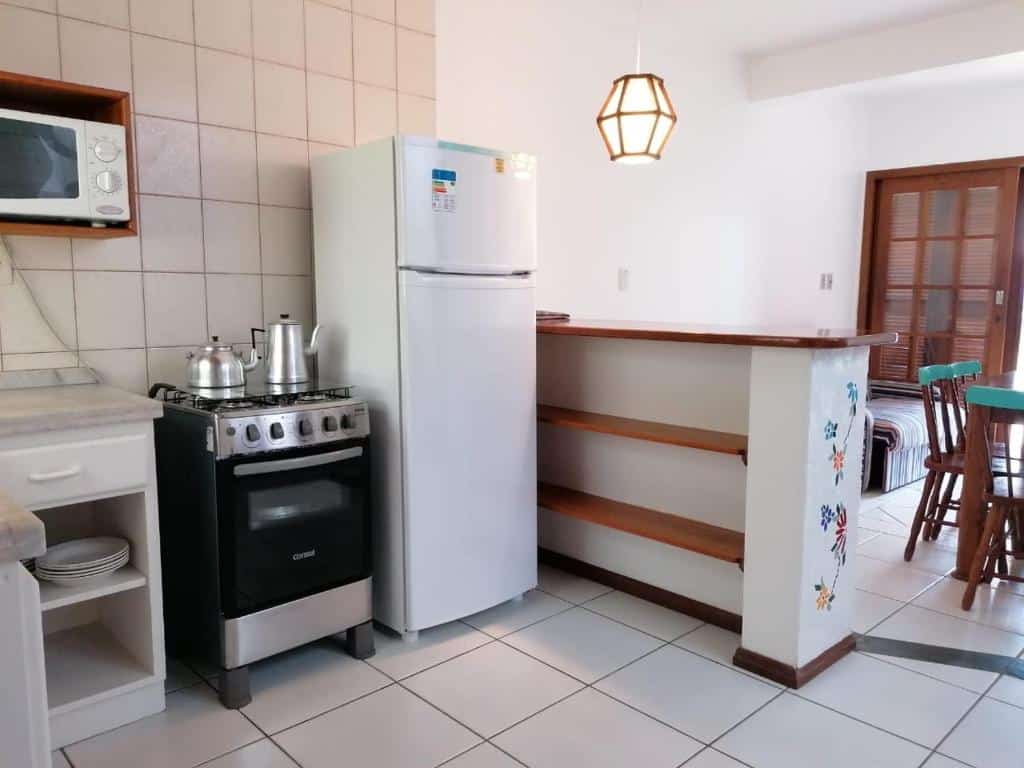Cozinha do Condominio Vila Lila. Ao lado esquerdo da imagem há um balcão com utensílios; em um suporte na parede acima fica o micro-ondas; ao lado da bancada está o fogão com chaleiras e, ao lado, a geladeira. Na lateral direita, uma bancada maior separa a cozinha da sala de estar, onde é possível ver um pedaço do sofá e da mesa de jantar.