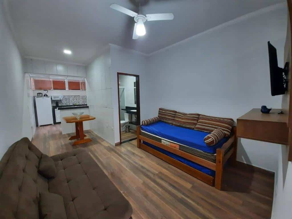 Parte do Condomínio Ville I que mostra um sofá na parede esquerda e um sofá-cama na parede direita e em outra parede uma tv. Ao fundo tem uma porta que dá acesso ao banheiro e do outro lado a cozinha. Imagem ilustrando post Airbnb em Maranduba.