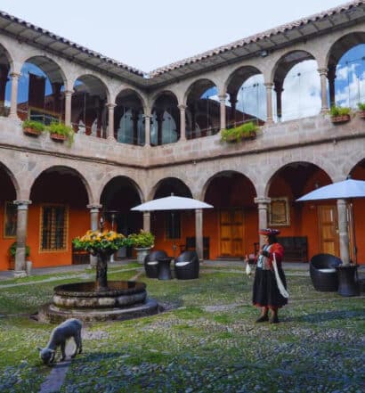 pátio aberto do hotel Costa del Sol Wyndham em Cusco com uma fonte de pedra ao centro e arcos de pedra nas paredes atrás. Há também um cordeinrinho branco e uma mulher com roupas típicas peruanas.