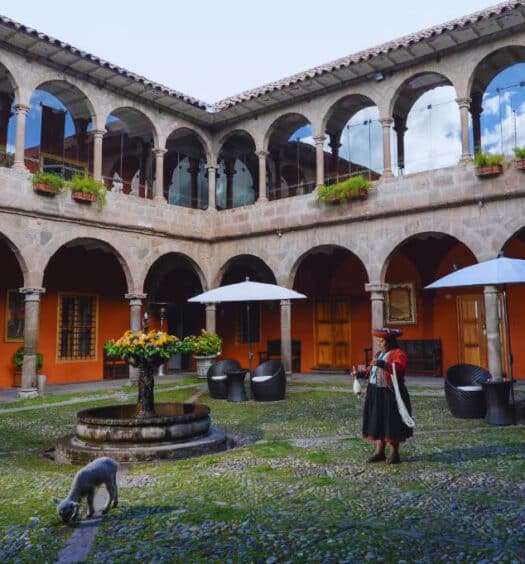 pátio aberto do hotel Costa del Sol Wyndham em Cusco com uma fonte de pedra ao centro e arcos de pedra nas paredes atrás. Há também um cordeinrinho branco e uma mulher com roupas típicas peruanas.