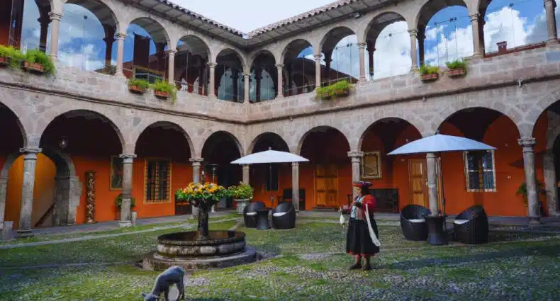 pátio aberto do hotel Costa del Sol Wyndham em Cusco com uma fonte de pedra ao centro e arcos de pedra nas paredes atrás. Há também um cordeinrinho branco e uma mulher com roupas típicas peruanas.