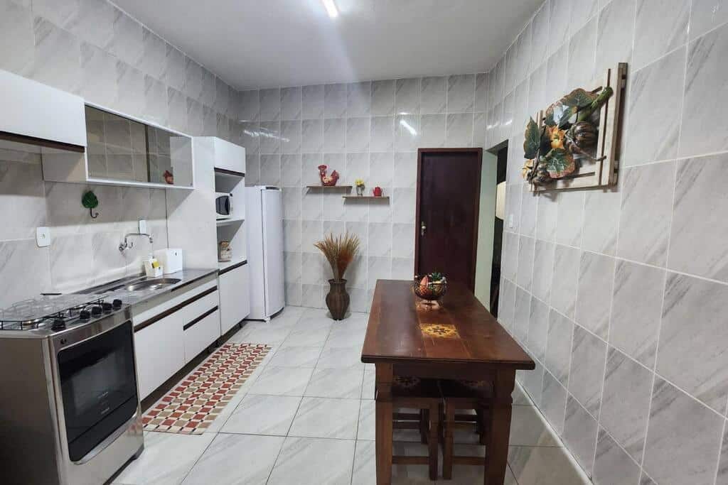 Cozinha da Casa dos Sinos do lado direito uma mesa de madeira com bancos e do lado esquerdo um fogão, uma pia, armário e geladeira.