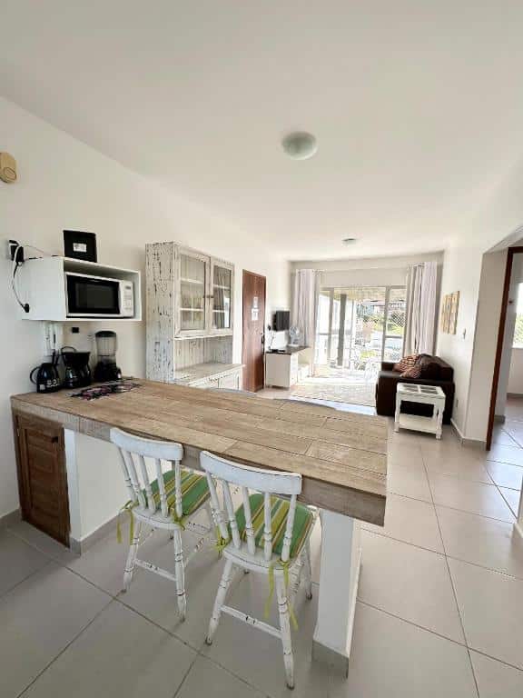 Cozinha e sala do airbnb Residencial Mansur. Na frente da imagem há uma bancada de madeira com duas cadeiras, um micro-ondas a esquerda e ao fundo está a sala de estar, que conta com sofá e televisão e uma varanda ainda mais ao fundo.