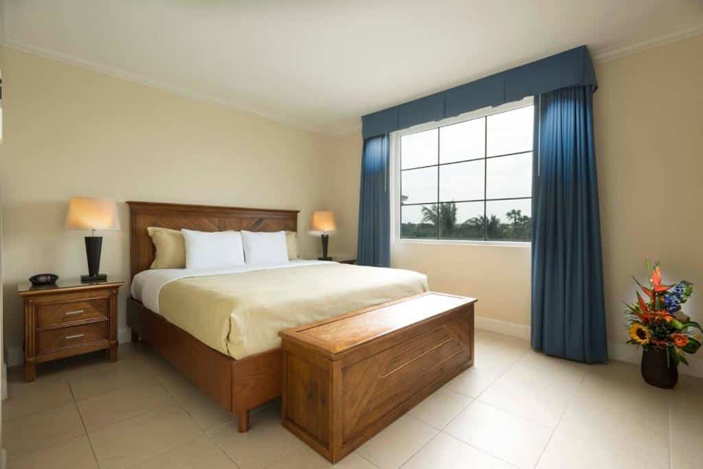 Quarto do All Inclusive - Divi Village Golf and Beach Resort. No meio uma cama de casal, na frente um baú, de cada lado uma cômoda com abajur. No fundo, uma janela com cortina.