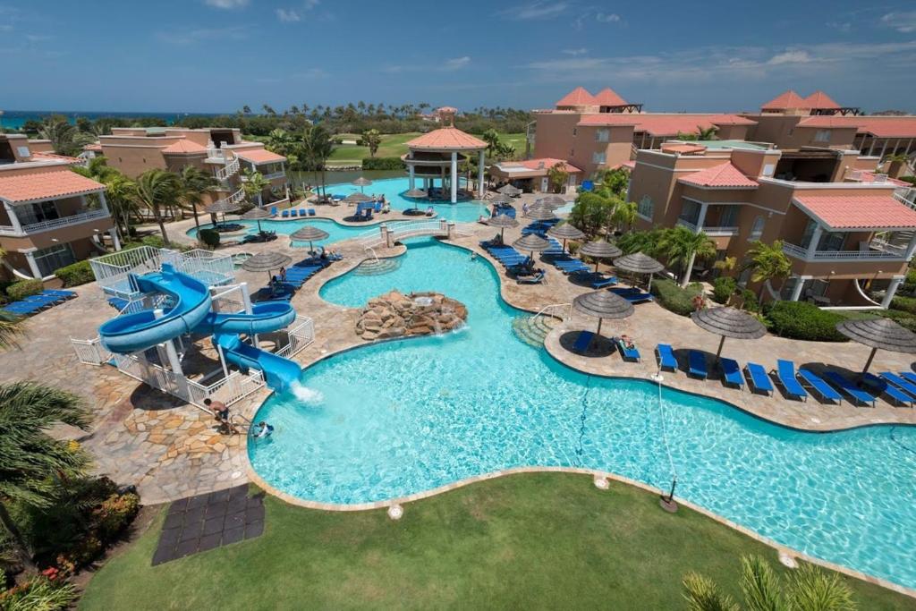 Vista de cima da área de lazer do All Inclusive - Divi Village Golf and Beach Resort. Uma piscina frande, com cadeiras e guarda-sóis ao redor. Do lado esquerdo um tobogã.