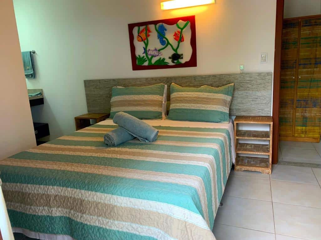 Foto do quarto em Duplex com hidromassagem - Praia do Forte, que ilustra o post de airbnb na Praia do Forte. No meio há uma cama de casal com toalhas em cima e mesa de cabeceira em seus dois lados. Um quadro enfeita a parede atrás da cama.