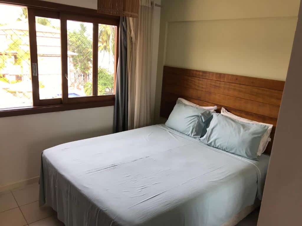 Foto do quarto em Enseada Praia do Forte, que ilustra o post de airbnb na Praia do Forte. A cama é box e de casal, atrás possui uma cabeceira de madeira. Do seu lado esquerdo há uma janela com rede.