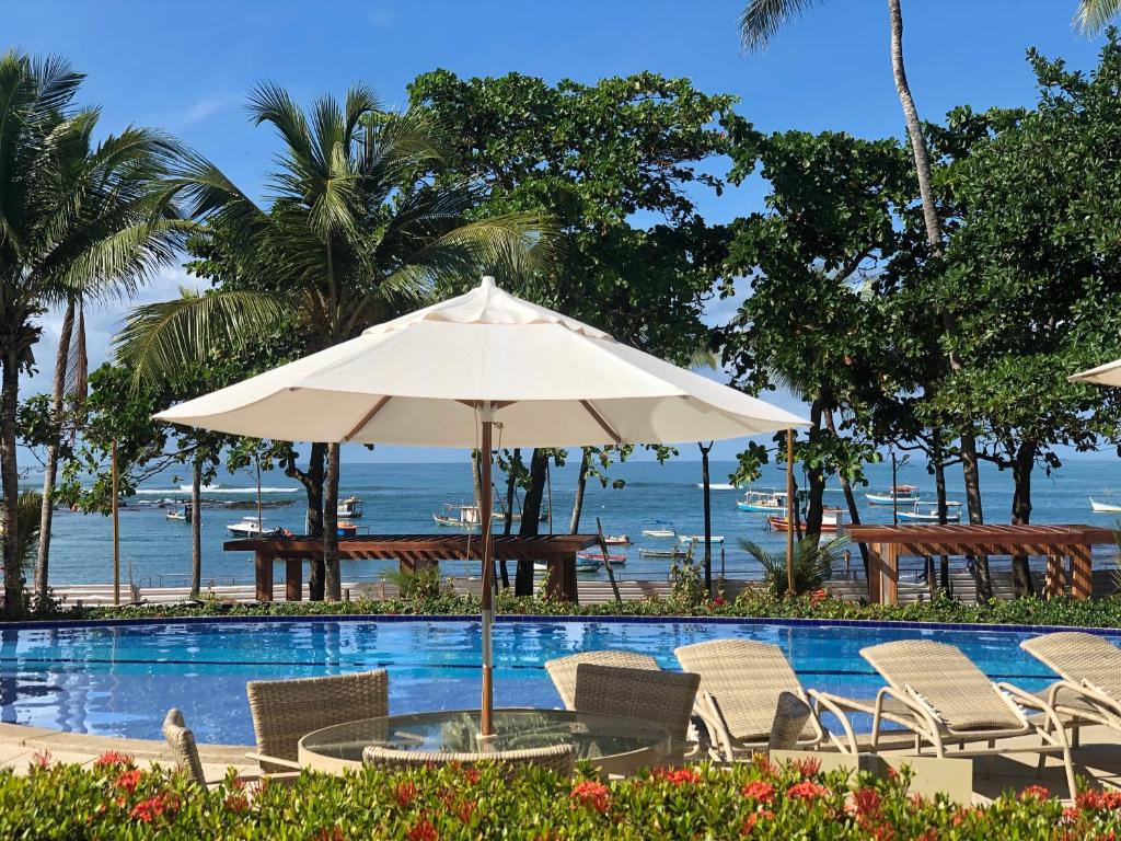 Piscina com espreguiçadeiras, guarda-sol e jardim na Enseada Praia do Forte. No fundo está o mar, que conta com vários barcos.