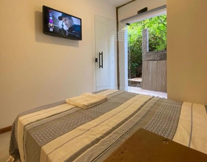 Quarto do Estúdio Vista Mar. Uma cama de casal está de frente para uma parede com uma tv. Ao lado da cama está uma porta de vidro, para representar airbnb na Praia Vermelha do Centro.