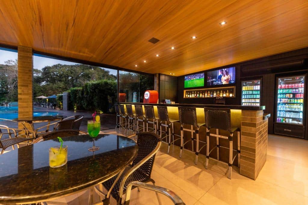 Parte do Falls Galli Hotel que mostra do lado direito o bar do hotel com uma bancada com várias banquetas e ao fundo geladeiras com bebidas, duas tv na parede e outra parte com bebidas. Do lado esquerdo tem algumas mesas redondas com cadeiras e ao fundo uma piscina ao ar livre. Imagem ilustrando post Hotéis em Foz do Iguaçu.