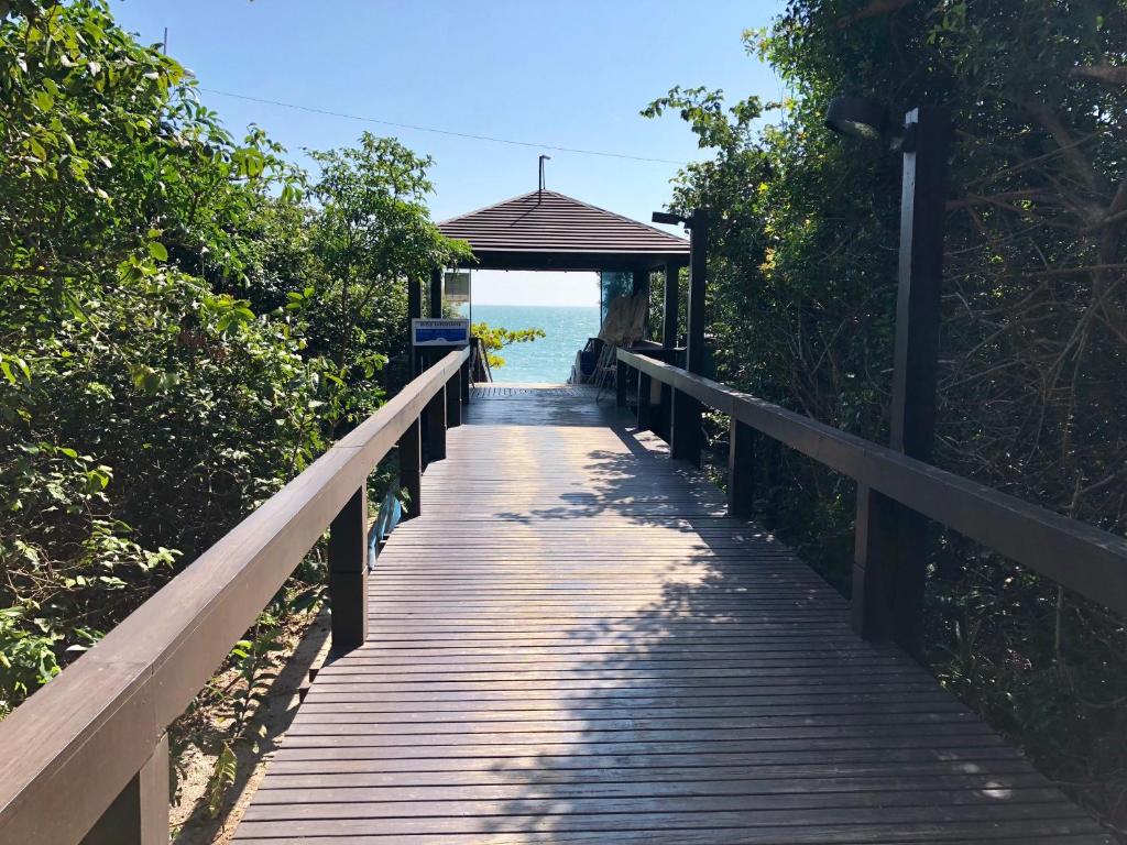 Área externa do airbnb Flat Partic Hotel Jurere Beach Village. Deck de madeira com passarela e corrimãos dos dois lados, cercado por uma área verde. Ao fundo é possível ver a praia.