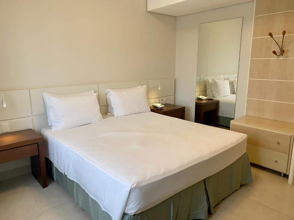 Quarto do airbnb Flat Partic Hotel Jurere Beach Village. No lado direito há um espelho de corpo inteiro, ao lado do espelho há um painel de madeira com um cabideiro preso a ele e duas gavetas, também de madeira, abaixo. No centro do quarto há uma cama de casal, dos dois lados da cama têm cômodas feitas em madeira, com gavetas e pequenas luzes presas a um painel que fica na parede atrás a cama. Imagem utilizada para ilustrar o post airbnb em Jurerê Internacional.