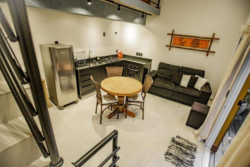 Cozinha de um dos Flats de Sumatra. No canto esquerdo está a geladeira, a pia e o fogão e forno. Ao lado do forno, na outra parede está encostado o sofá. No meio do ambiente há uma mesa redonda de três lugares. No canto é possível ver o pedaço de uma escada. Esse é um dos airbnb em Boiçucanga.
