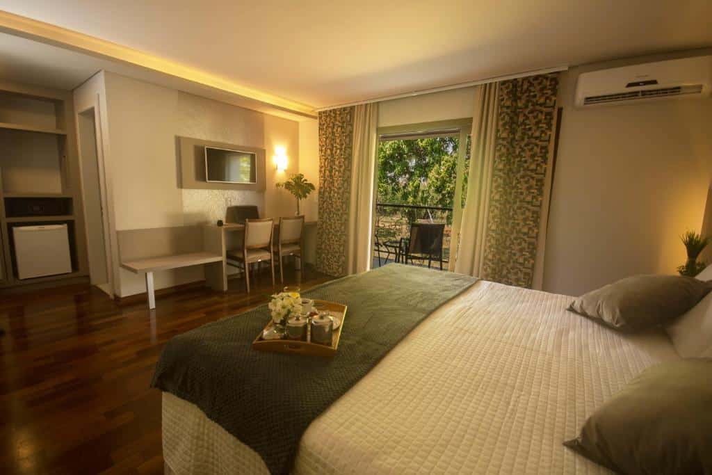 Foto do quarto do Grand Carimã Resort & Convention Center, ilustrando o post sobre Onde ficar em Foz do Iguaçu. A cama box de casal está na direita, e na frente dela há uma mesa com duas cadeiras e uma TV. Mais ao canto no fundo à esquerda há uma estante com frigobar. Já do lado direito há uma varanda com vista para o jardim.