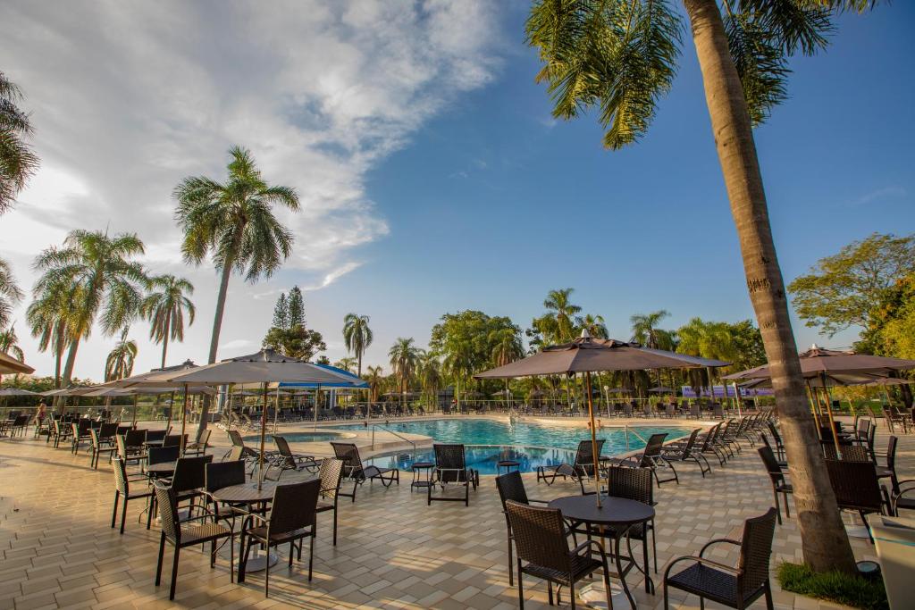 Foto do Grand Carimã Resort & Convention Center, mostrando um dia na piscina do hotel. Ao redor dela há muitas mesas com cadeira e guarda-sol. Além disso, há árvores pelo espaço.