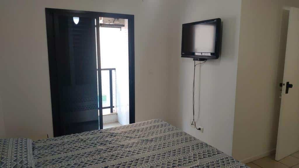 Imagem do quarto do Guarujá Pitangueiras Flat Capitania Varam, ilustrando o post sobre airbnb em Guarujá. Há uma cama de casal, que vemos de lado. Na frente dela há uma parede com TV fixa, e ao seu lado há uma porta com varanda.
