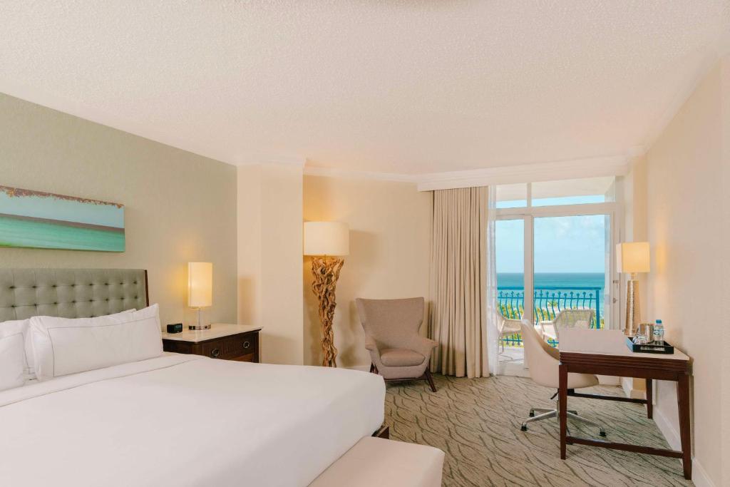 Quarto do Hilton Aruba Caribbean Resort & Casino. Do lado esquerdo uma cama de casal, e um acento na frente, do lado da cama dois abajures, uma cômoda e uma poltrona. Do lado direito uma mesa de trabalho, um abajur e a porta da varanda com vista para o mar.