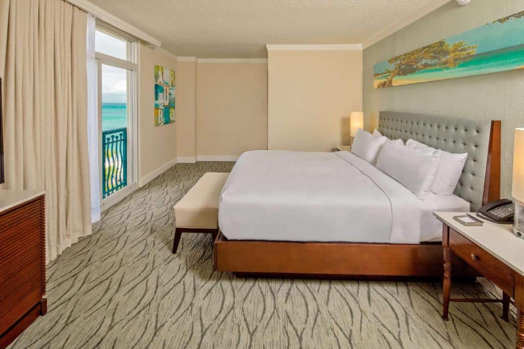 Quarto do Hilton Caribbean Resort & Casino. Uma cama de casal do lado direito com uma cômoda de cada lado e um abajur, na frente um acento. Do lado esquerdo uma cômoda e a cortina com a vista do mar. Foto para ilustrar post de onde ficar em Aruba.