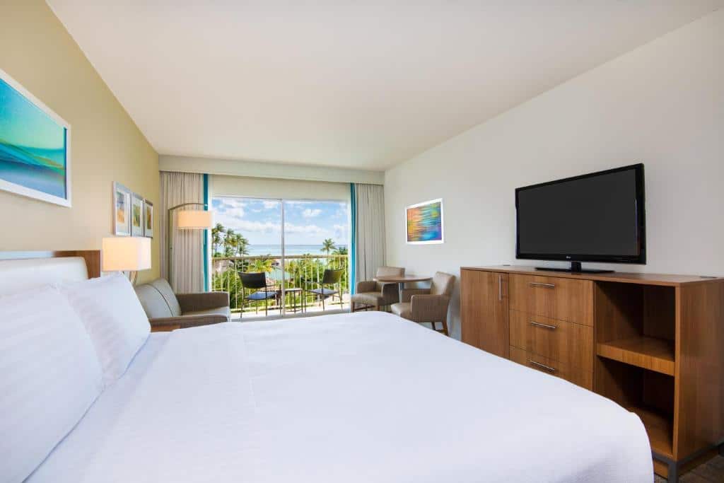 Quarto do Holiday Inn Resort Aruba - Beach Resort & Casino. Do lado direito uma cômoda com televisão, duas cadeiras e uma mesa. Do lado esquerdo a cama de casal, dois abajures e um sofá. No fundo a varanda com vista do mar.