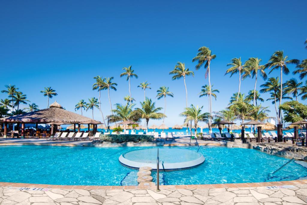 Área de lazer do Holiday Inn Resort Aruba - Beach Resort & Casino. Uma piscina no centro, atrás cadeiras de tomar sol, guarda-sol e palmeiras, no fundo a praia.