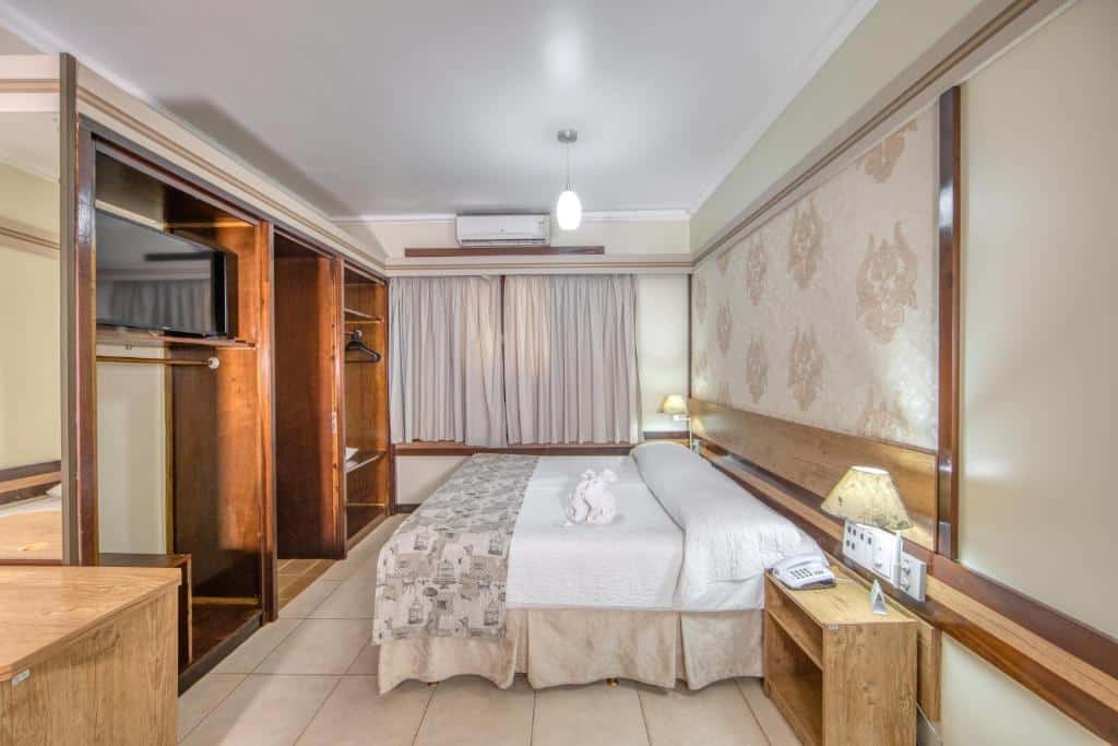 Foto do quarto do Hotel Colonial Iguaçu, ilustrando o post sobre Onde ficar em Foz do Iguaçu. Há uma cama box de casal na direita, e na frente há armários e uma TV. No fundo há uma janela e um ar-condicionado.