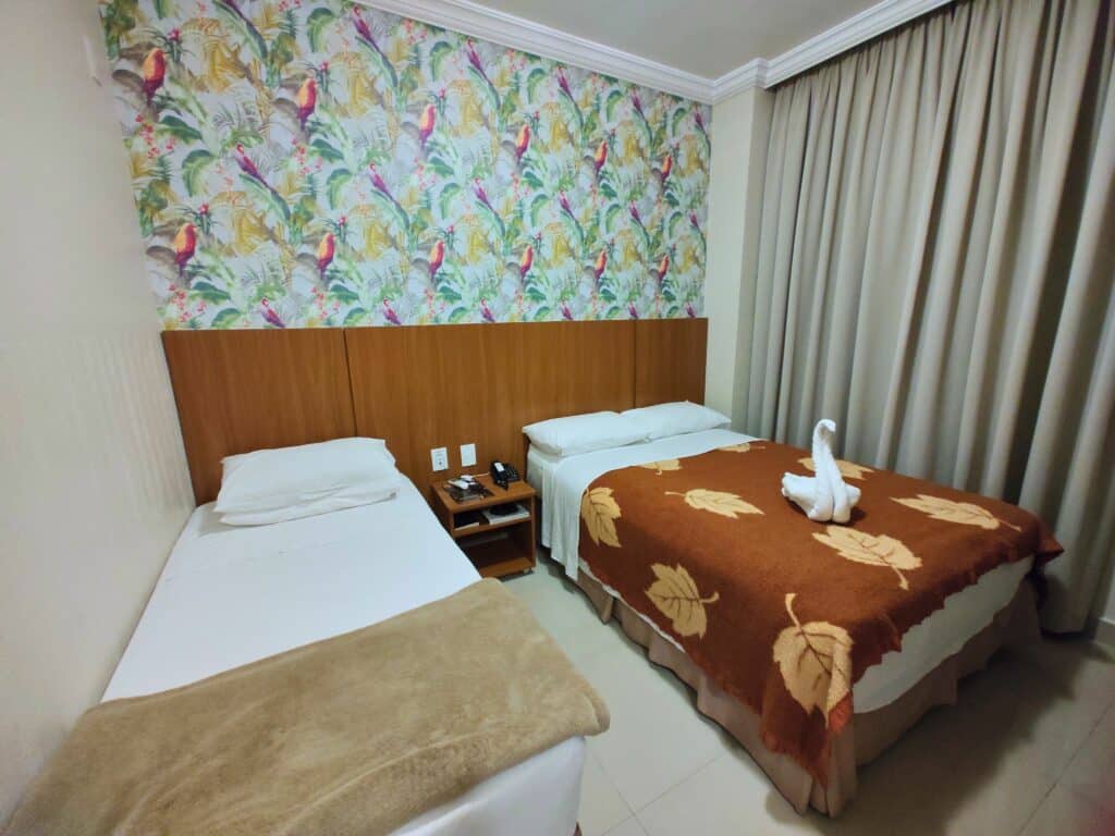 Quarto do Del Rey Quality Hotel com duas camas, uma cama de solteiro e do lado uma cama de casal. Separando as camas tem uma mesa de cabeceira e atrás uma cabeceira de madeira com parede colorida. Do lado da cama de casal tem uma cortina grande.