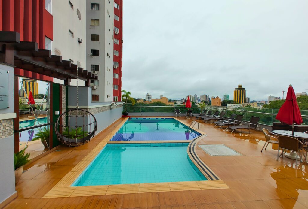 Parte do Del Rey Quality Hotel que mostra a piscina do hotel em volta de um piso de madeira. Do lado esquerdo tem a construção do prédio e uma cobertura com uma balança. Já do lado direito tem algumas espreguiçadeiras e mesas com guarda-sóis e uma vista para os prédios e árvores da cidade. Imagem ilustrando Hotéis em Foz do Iguaçu.