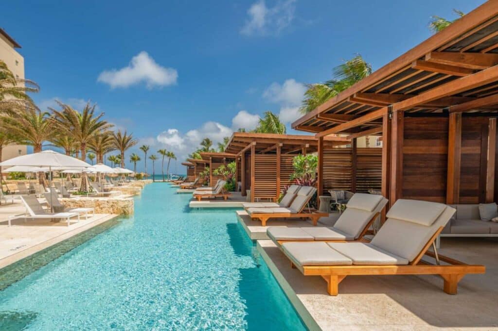 Uma das piscinas do Hyatt Regency Aruba Resort & Casino. Ela é mais fina e comprida e passa no meio de uma área com várias espreguiçadeiras e cabanas de madeiras. Ao fundo está o mar.