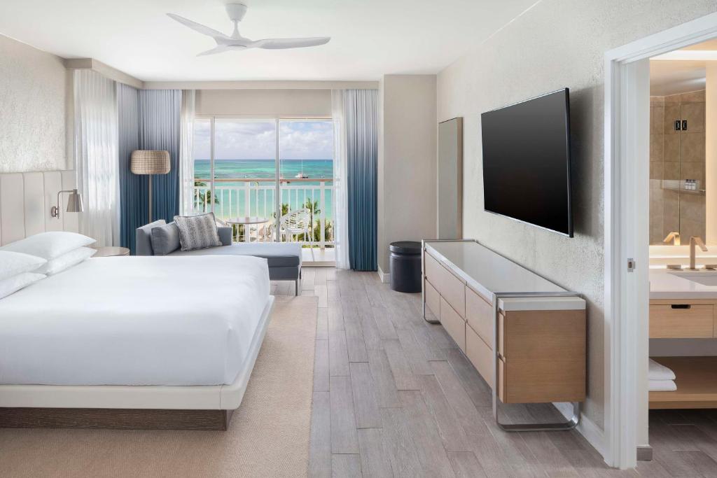 Quarto do Hyatt Regency Resort & Casino. Do lado direito uma raque uma televisão e a porta do banheiro. Do lado esquerdo a cama de casal, no fundo um mini sofá com luminária e varanda com vista do mar. Foto para ilustrar post de onde ficar em Aruba.