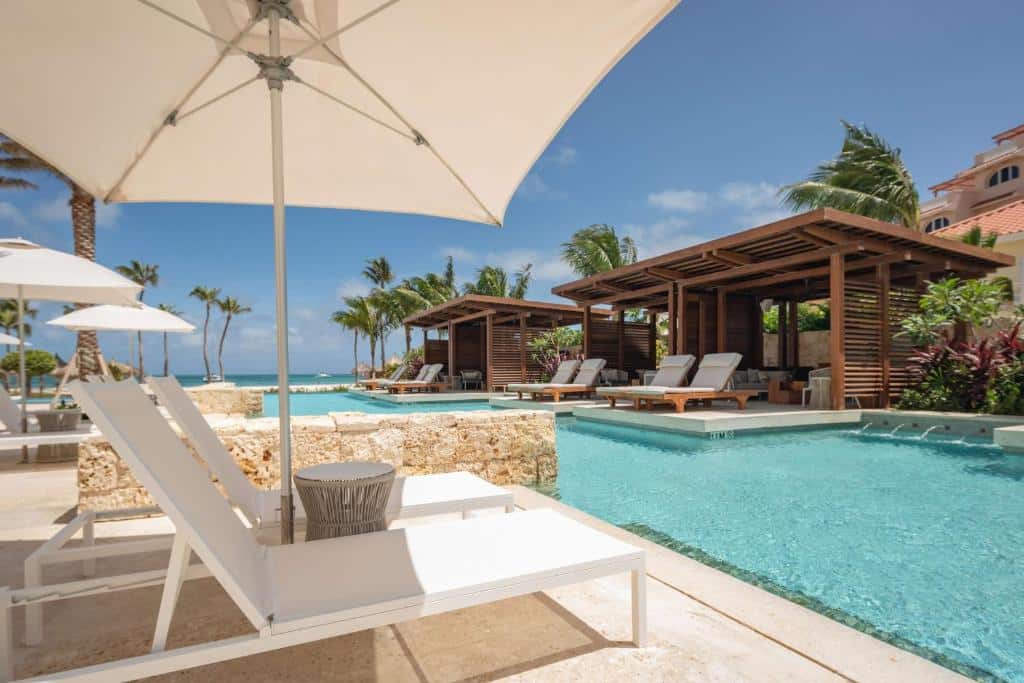 Área externa do Hyatt Regency Aruba Resort & Casino. Uma piscina no meio, de cada lado cadeiras de tomar sol, guarda-sóis, coberturas e no fundo a praia.