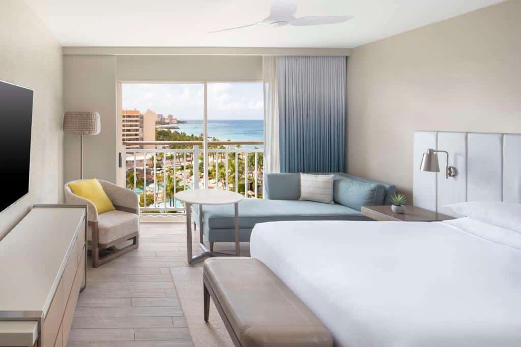 Quarto do Hyatt Regency Aruba Resort & Casino. Do lado direito uma cama de casal com um banco, uma mesa de apoio e uma luminária, um sofá e uma mesa de centro. Do lado esquerdo uma raque, televisão, poltrona e um abajur. No fundo a varanda.