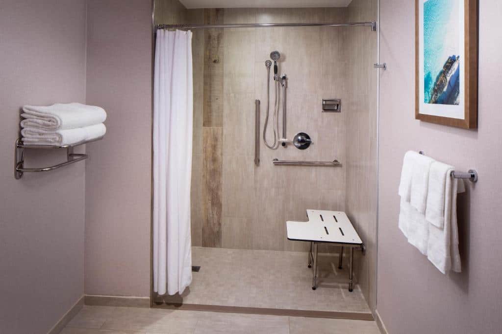 Banheiro do Hyatt Place Aruba Airport. Do lado direito toalhas penduradas, do lado esquerdo toalhas dobradas. No fundo o box com cadeira, mangueira do chuveiro e barras de apoio.