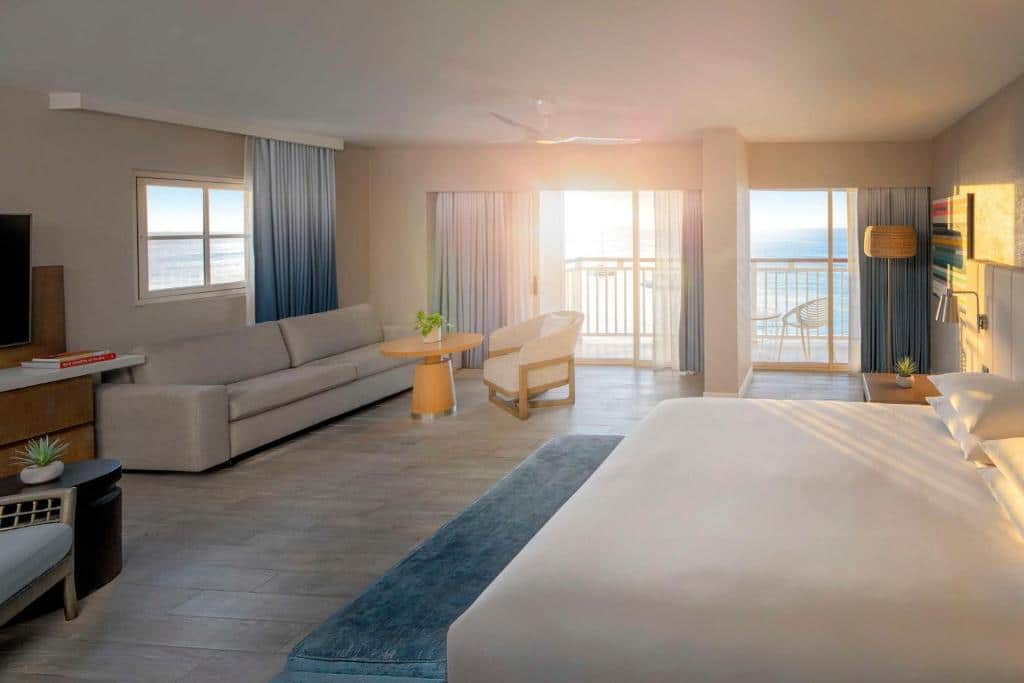 Quarto do Hyatt Regency Aruba Resort & Casino. Uma cama de casal está no meio, de frente para uma tv e um móvel. Ao fundo, na parede de frente para a cama, está um frande sofá, uma mesinha de centro e uma cadeira. Ao lado deles está a porta para a varanda com vista do mar. Este é um dos hotéis de luxo em Aruba.