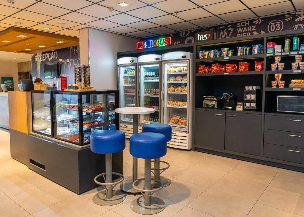 Parte do ibis budget Foz do Iguaçu que mostra uma parte de alimentos, com um armário com guloseimas e maquinas. No espaço há também três geladeiras, um balcão com comidas e uma mesinha redonda com três banquetas. Imagem ilustrando post Hotéis em Foz do Iguaçu.