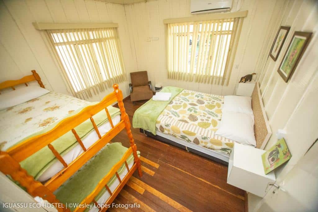 Foto do quarto do Ibis Foz do Iguaçu, ilustrando o post sobre Onde ficar em Foz do Iguaçu. Mostra de cima uma cama box de casal na direita e uma beliche na esquerda. Há duas janelas no quarto, ar-condicionado, uma poltrona e quadros decorando.