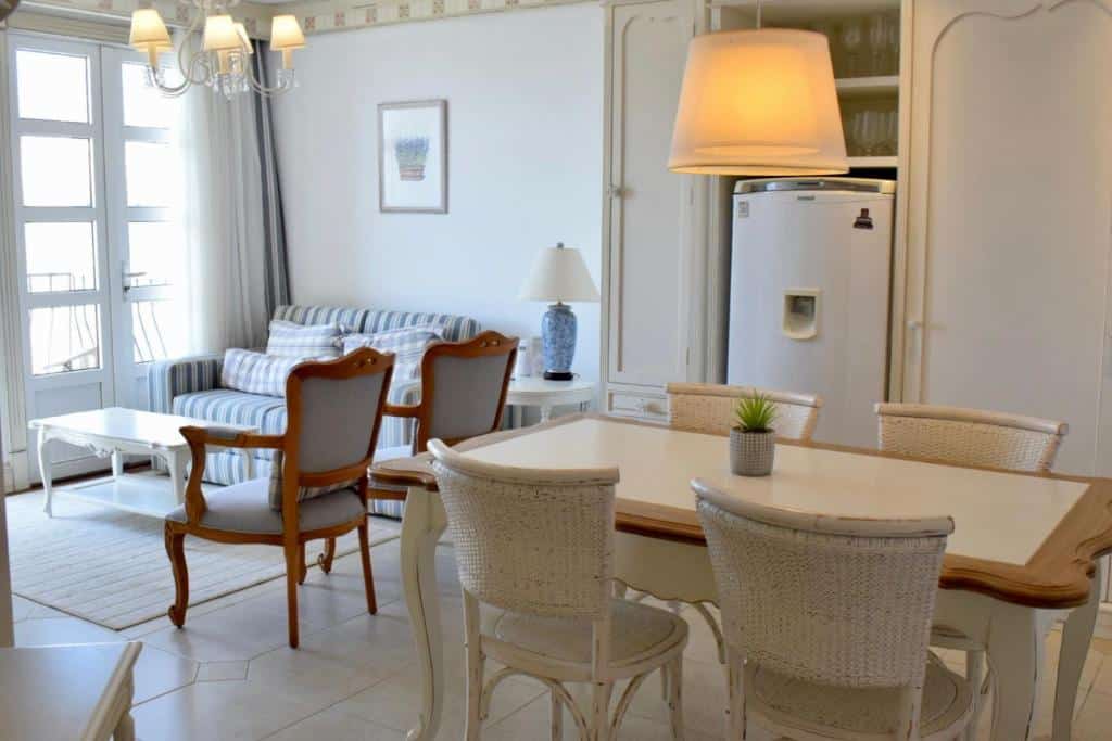 Sala de estar do airbnb Il Campanario - Cobertura. No lado direito da imagem há uma mesa com quatro lugares, na parede a frente da mesa há um armário e uma geladeira ao lado. No lado esquerdo ao fundo é possível ver a sala, que têm duas cadeiras, um sofá e uma porta com cortinas.