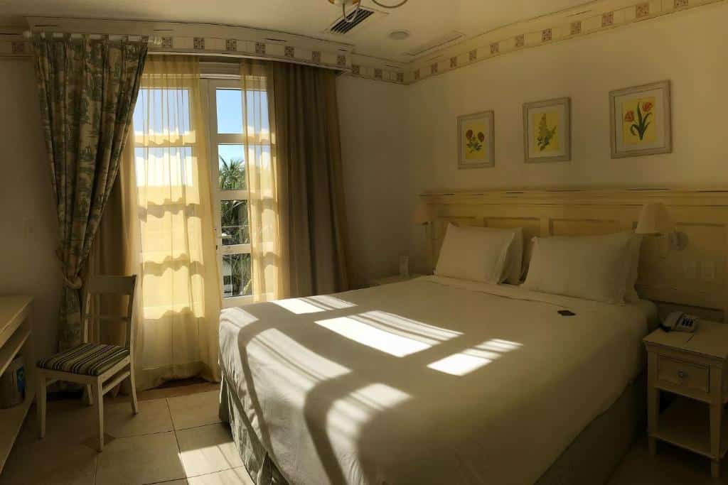 Quarto do airbnb IL Campanario é Destino Floripa. No centro do quarto há uma cama de casal, com cômodas dos dois lados e abajures presos ao painel da parede. Em frente a cama há uma cadeira. Ao fundo há uma porta com duas cortinas, que vai para a parte externa do quarto. Imagem utilizada para ilustrar o post airbnb em Jurerê Internacional.